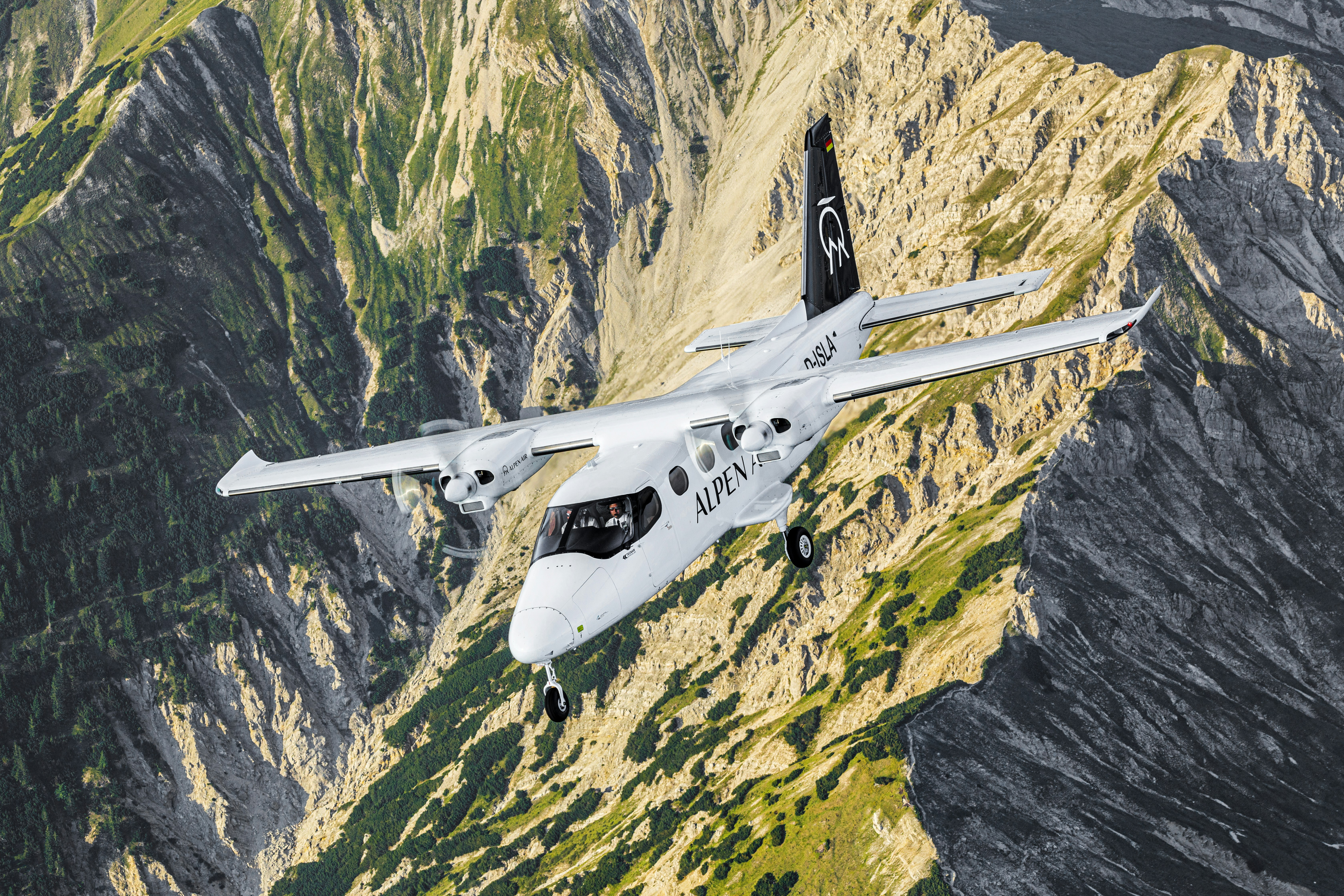 The white Alpen Air´s Tecnam P2012 Traveller airplane above the Bavarian landscape with rocky mountains in the Alps. Two pilots can be seen in the cockpit.